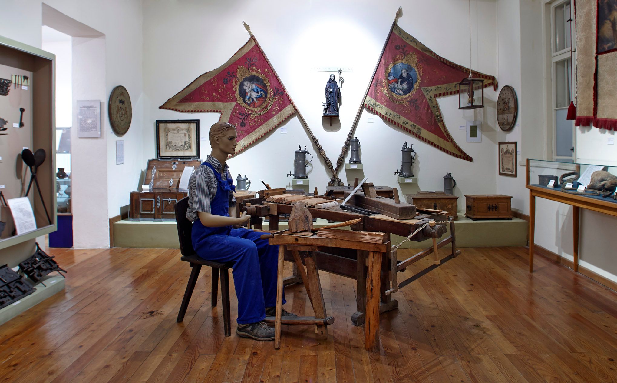 Interior view of a museum with a mannequin in work clothes at a wooden table, surrounded by historical objects and flags.