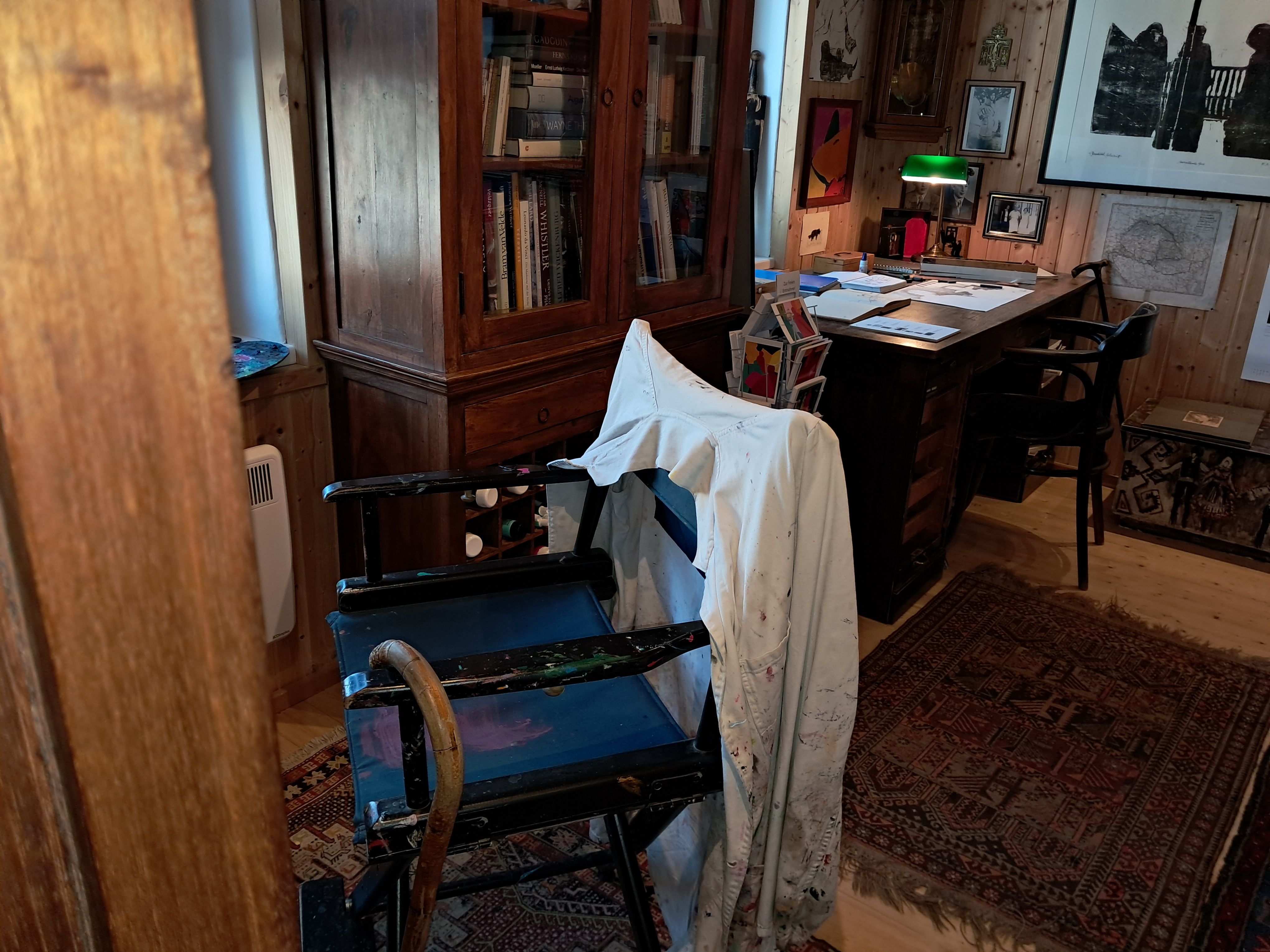 An artist's studio with a painted chair, a desk covered with papers, and a bookshelf in the background.