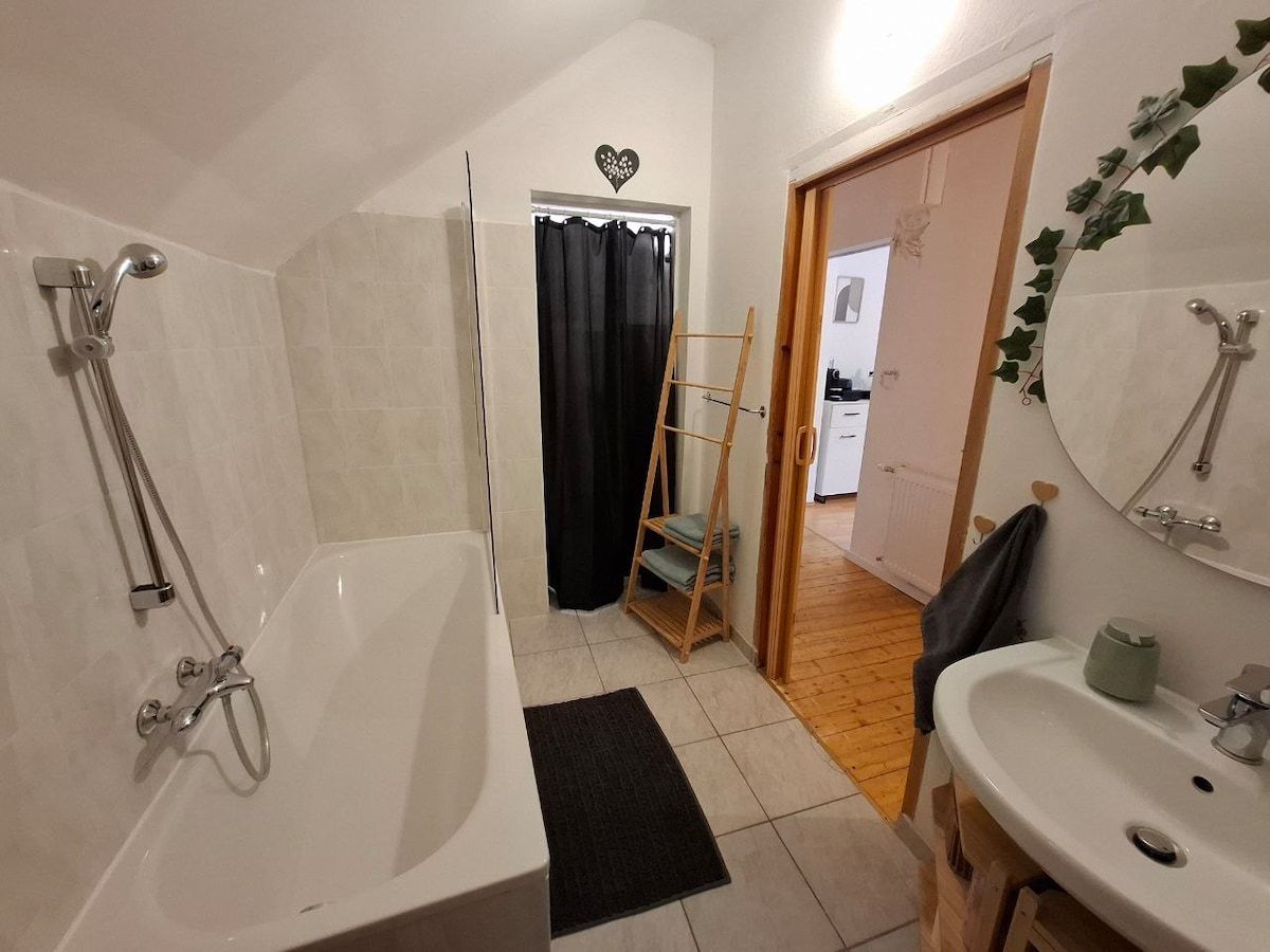 Ein Badezimmer mit Badewanne, Waschbecken und einem Handtuchhalter. Eine Tür führt zu einem angrenzenden Raum mit Holzboden.