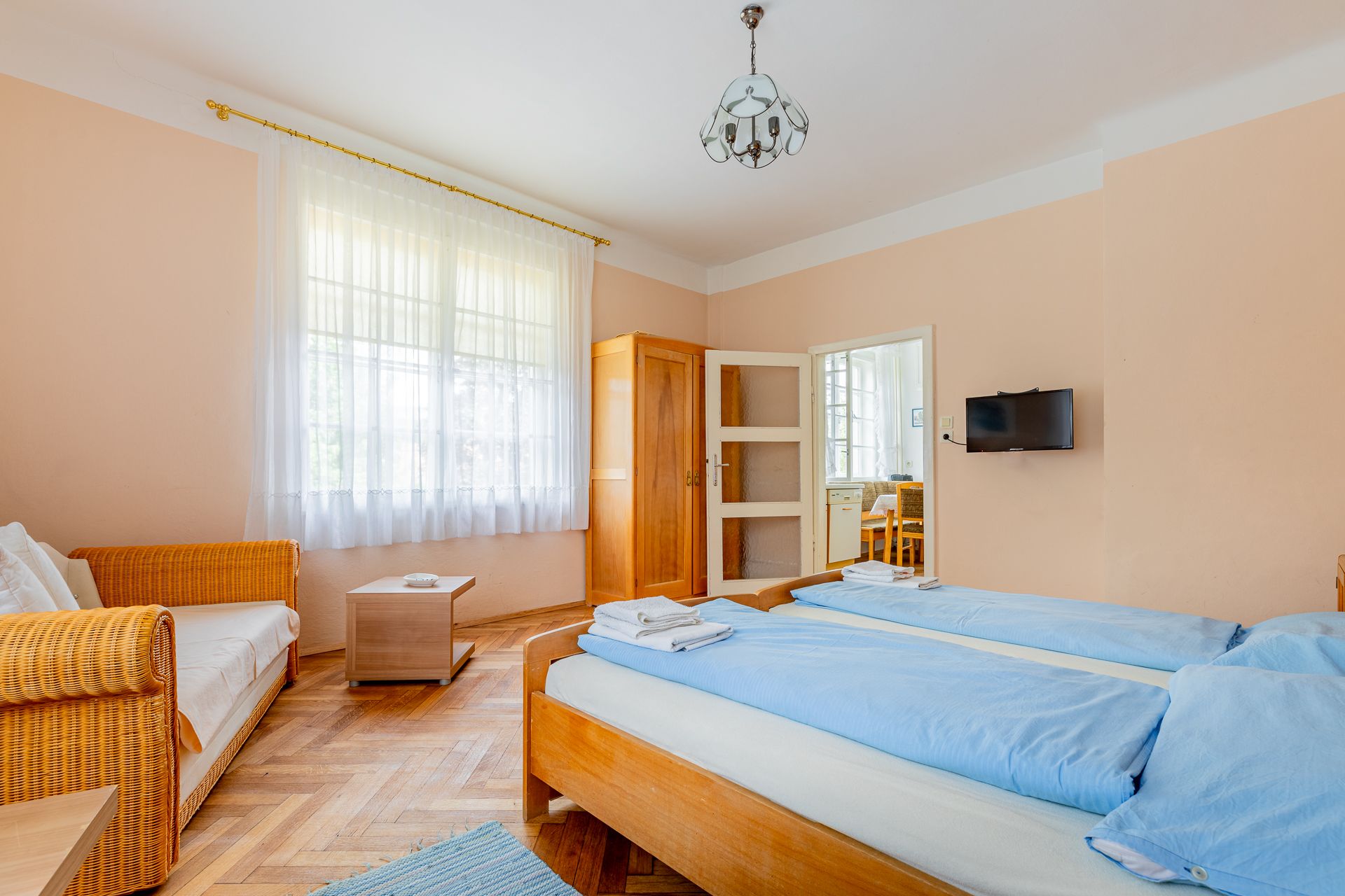 Ein gemütliches Schlafzimmer mit zwei Einzelbetten, einem Sofa, einem Schrank und einem kleinen Tisch. Ein Fenster mit Vorhängen lässt Tageslicht herein.