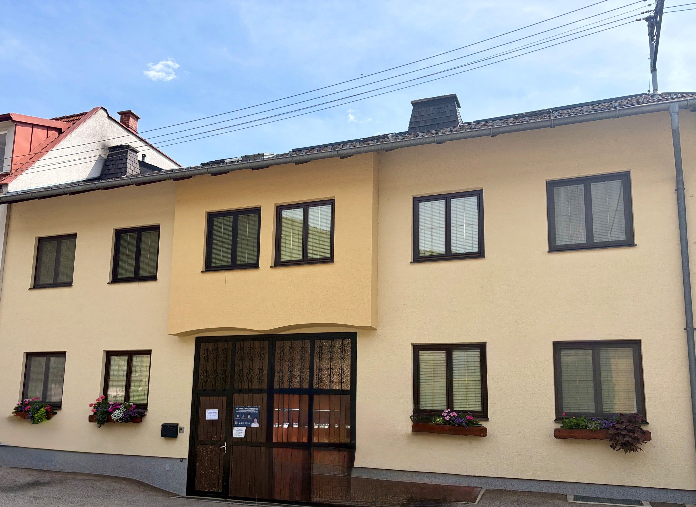 Fassade eines zweistöckigen Gebäudes mit gelber Wand und mehreren Fenstern. Blumenkästen schmücken einige Fenster. Der Himmel ist blau mit wenigen Wolken.