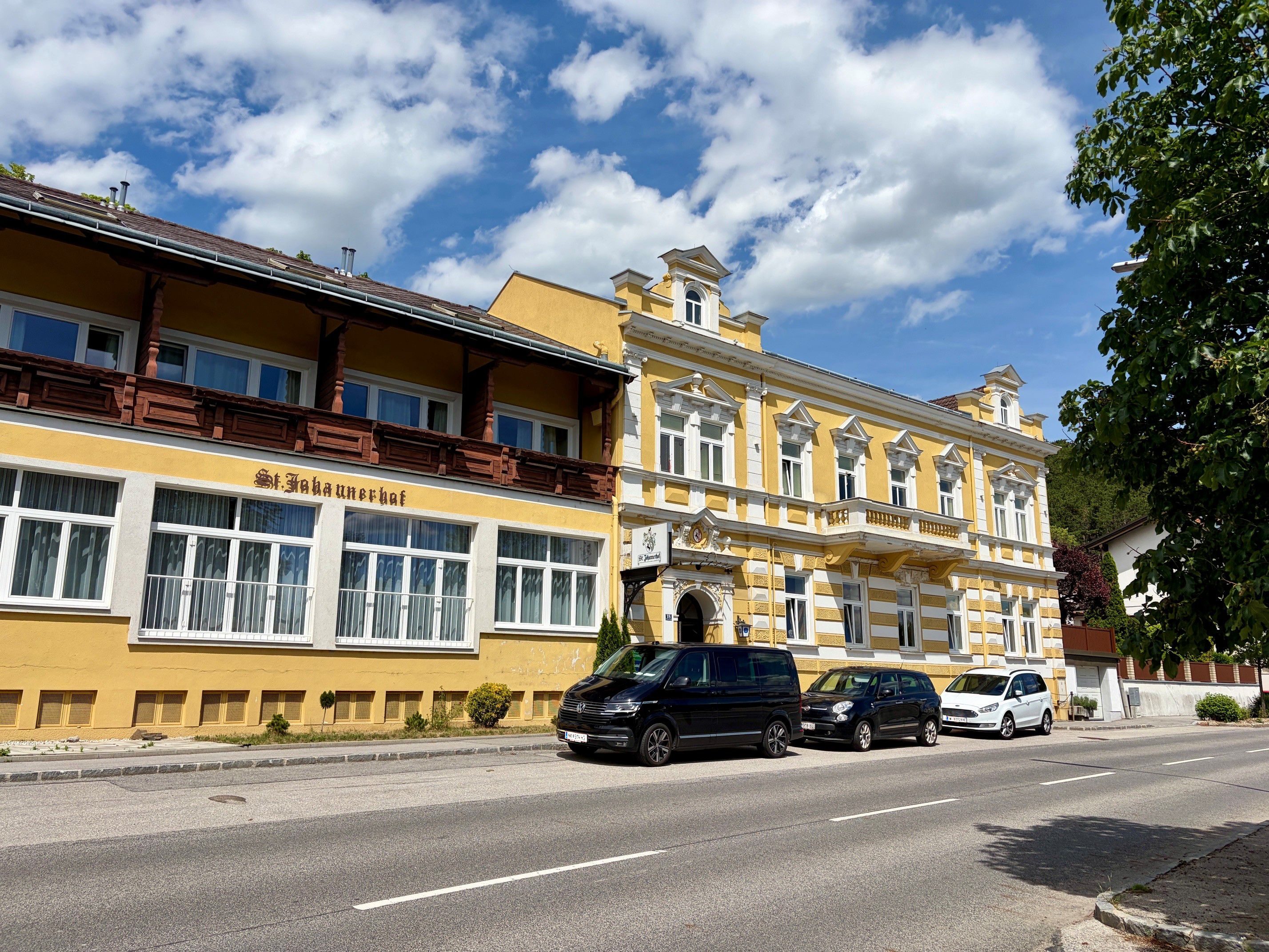 Gelbes Gebäude mit der Aufschrift 'St. Johannerhof' an einer Straße, umgeben von geparkten Autos und Bäumen.