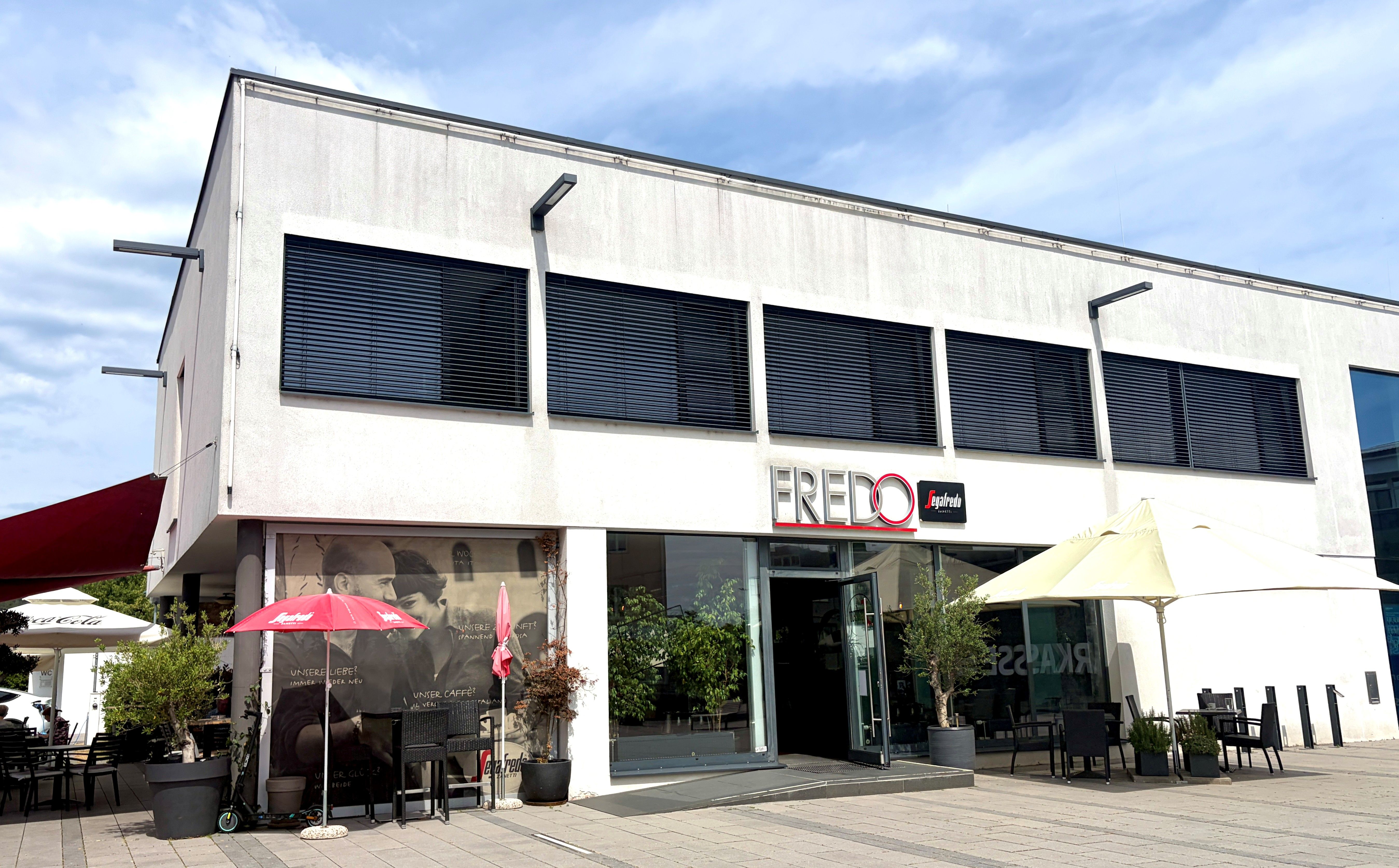 Exterior view of a modern café called Fredo, equipped with umbrellas and outdoor seating.