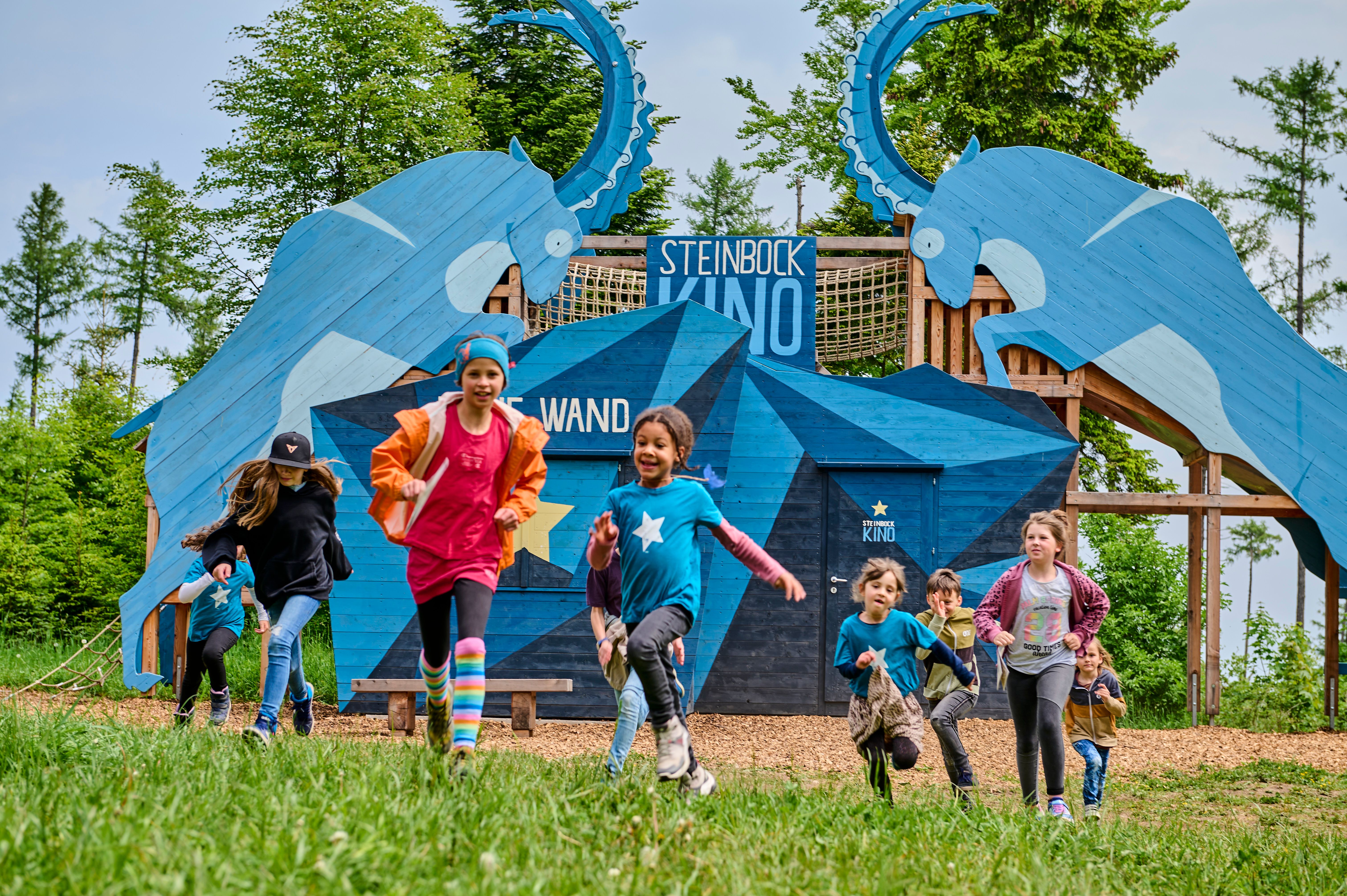Children run in front of a blue wooden construction with ibex motifs and the inscription 'Steinbock Kino'.