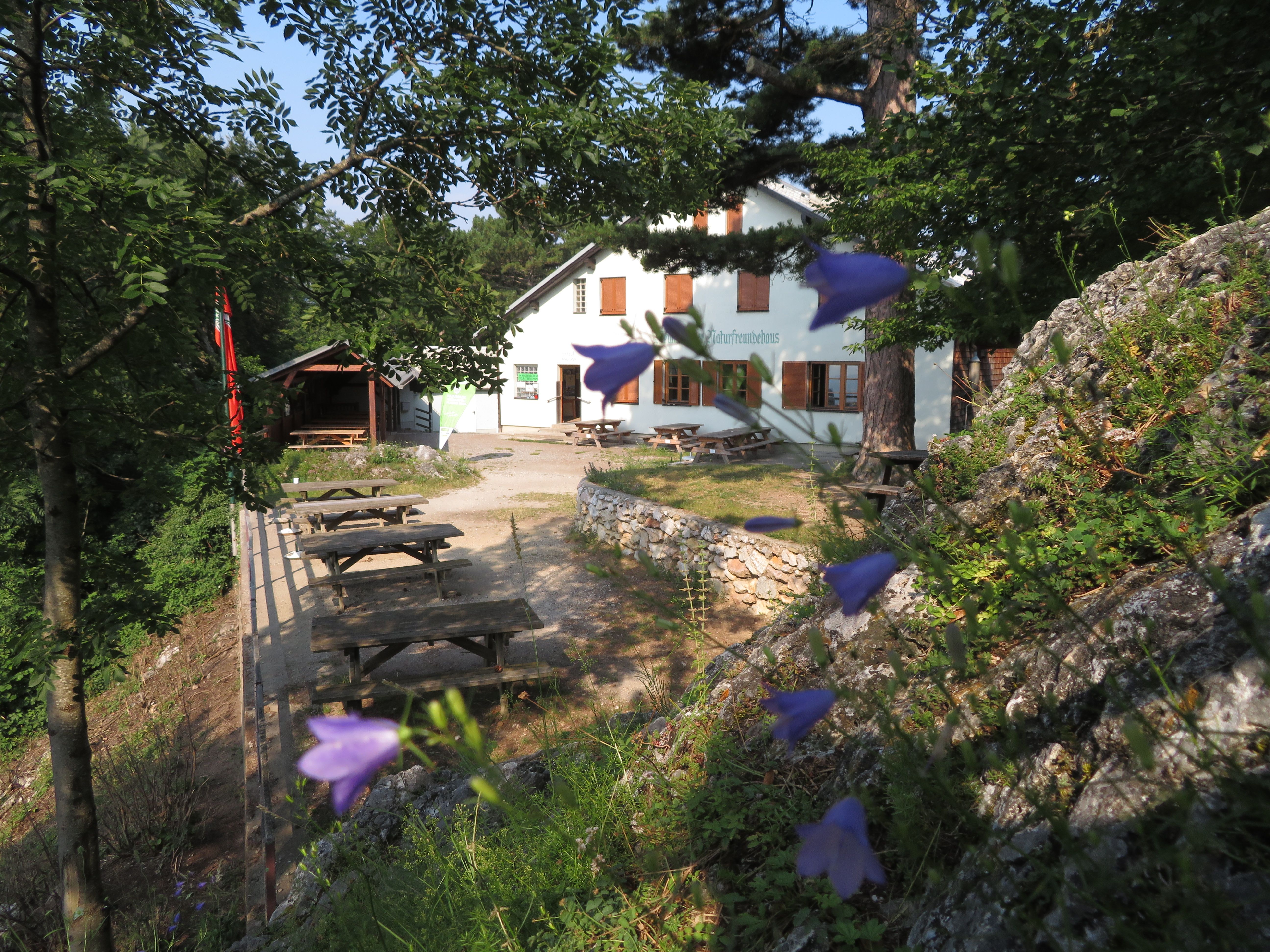 Ein weißes Gebäude mit der Aufschrift 'Naturfreundehaus' umgeben von Bäumen und Blumen. Im Vordergrund sind Holzbänke und Tische zu sehen.