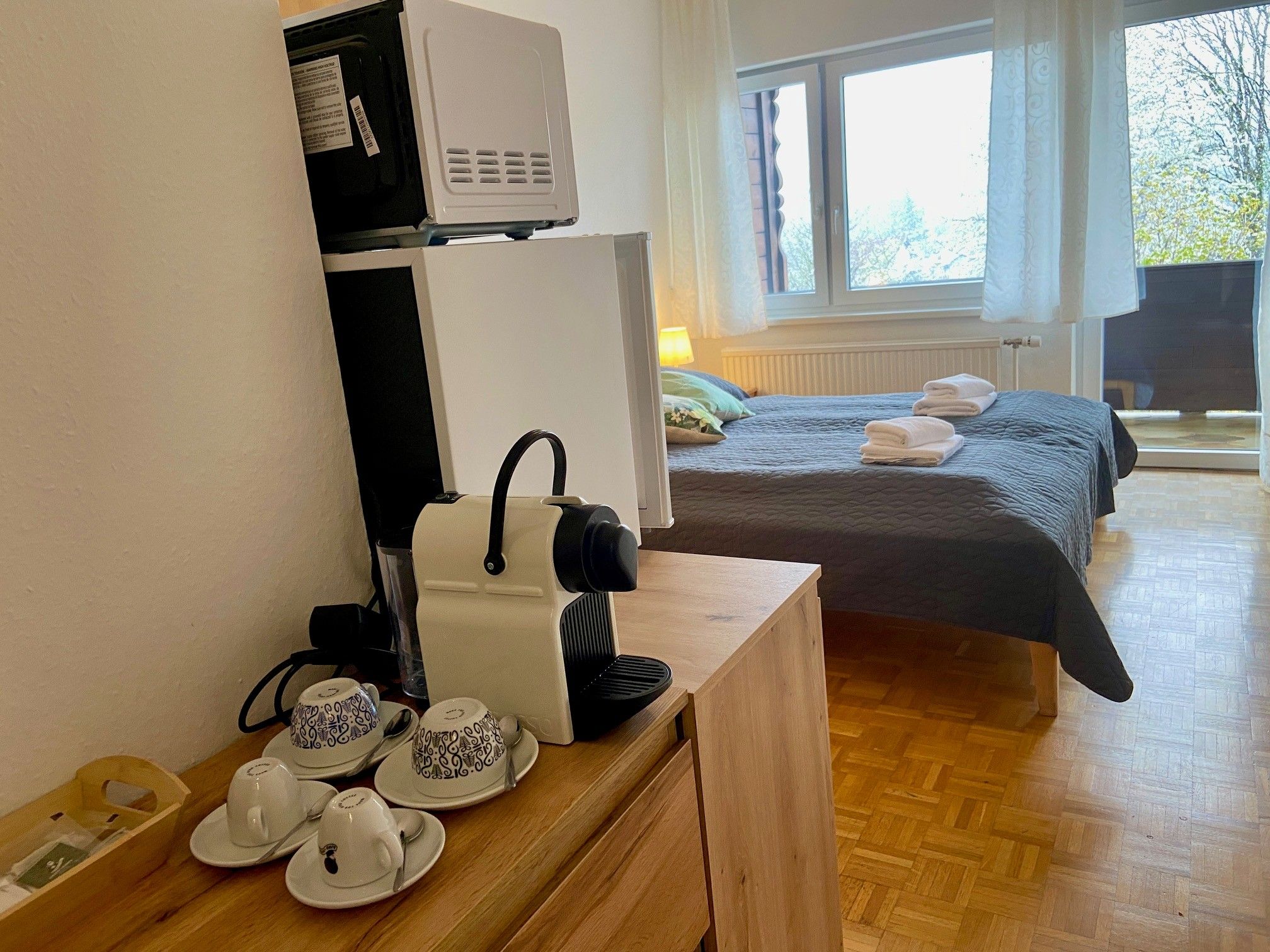 Ein Hotelzimmer mit einer Kaffeemaschine auf einem Tisch, daneben Tassen. Im Hintergrund ein Bett mit Handtüchern und ein Fenster mit Blick nach draußen.