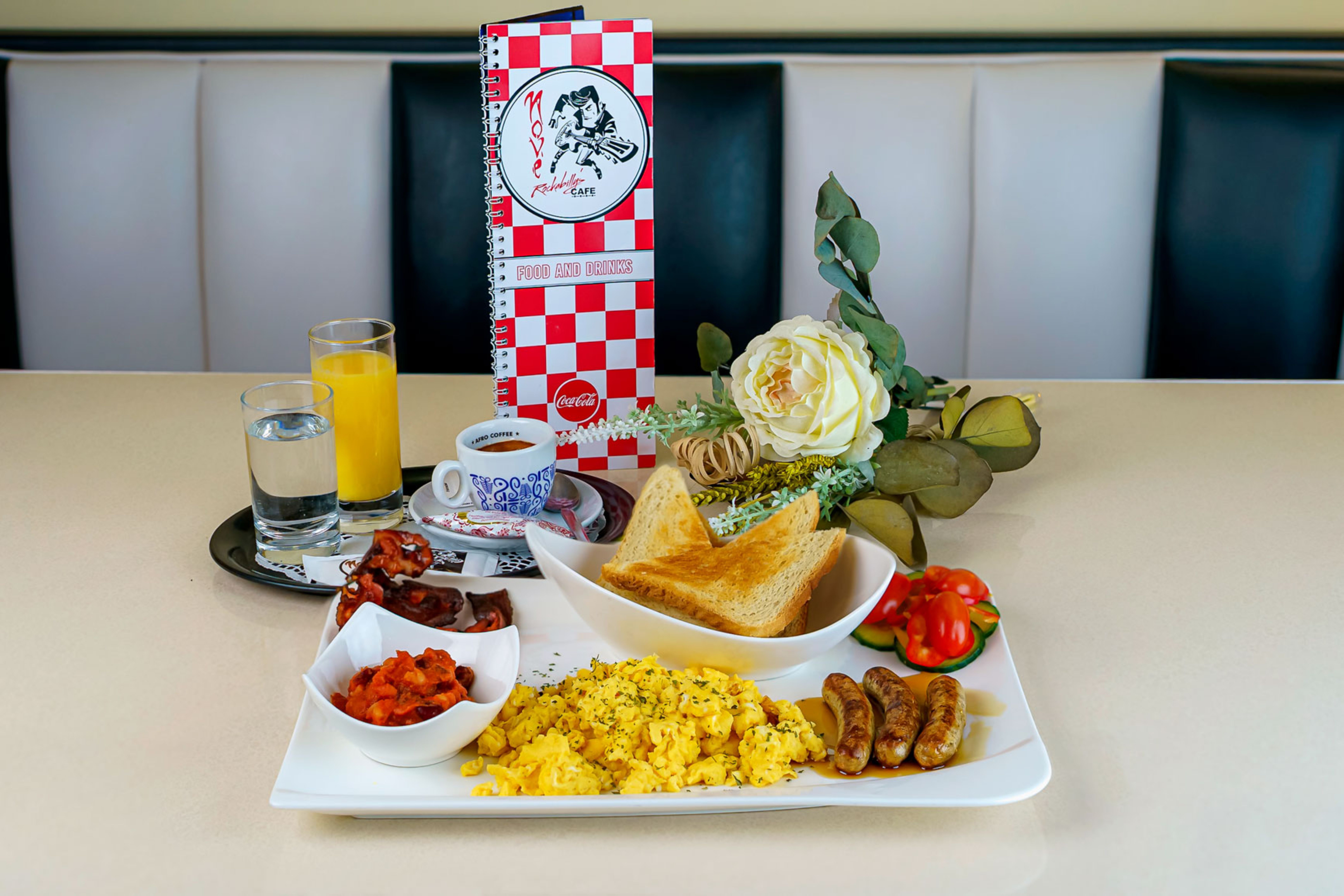 Frühstücksteller mit Rührei, Würstchen, Toast und Beilagen auf einem Tisch im Nové Rockabilly's Cafe.