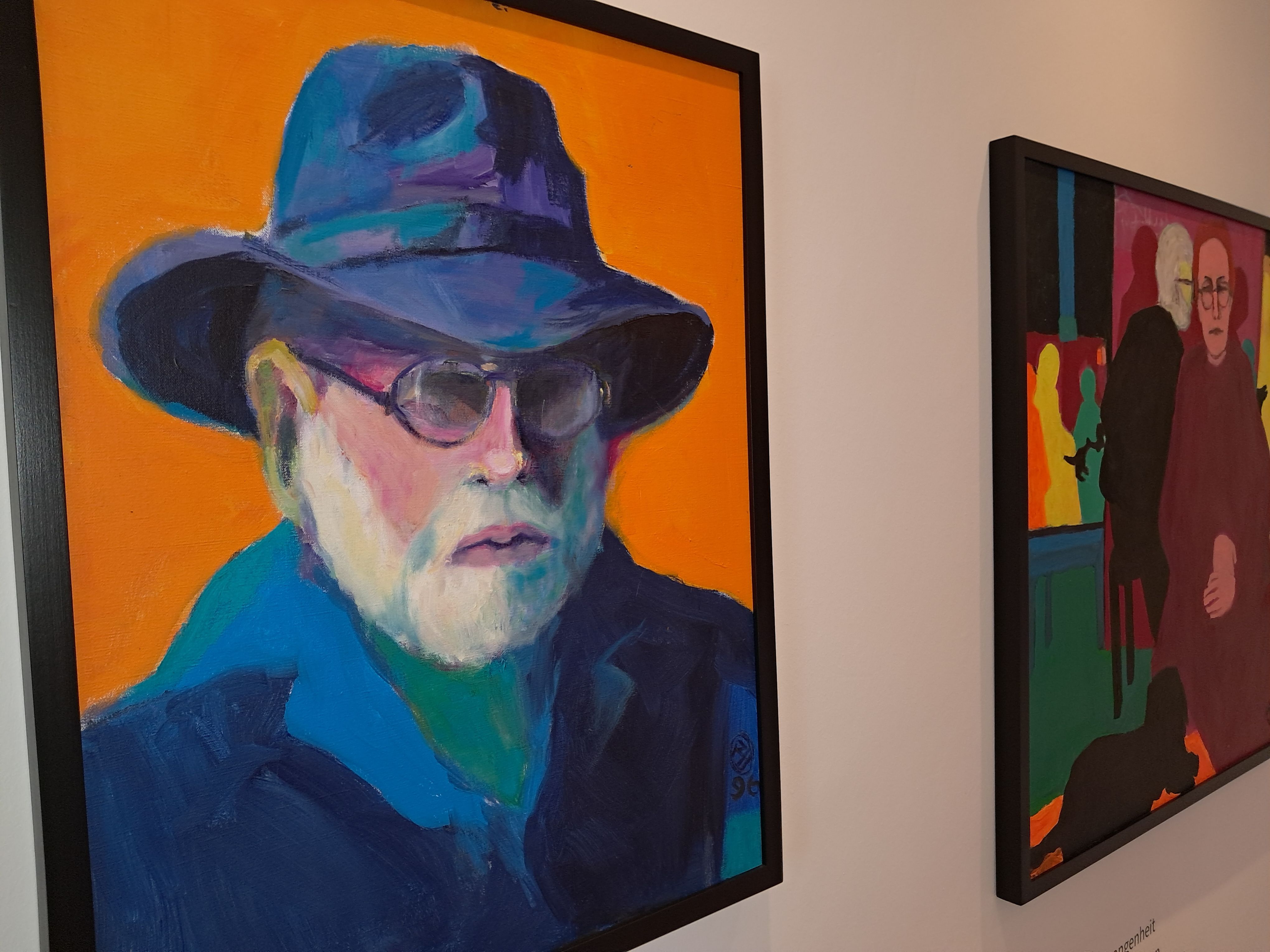 Two colorful paintings in a museum; one depicts a man wearing a hat and sunglasses, and the other depicts a group of people.