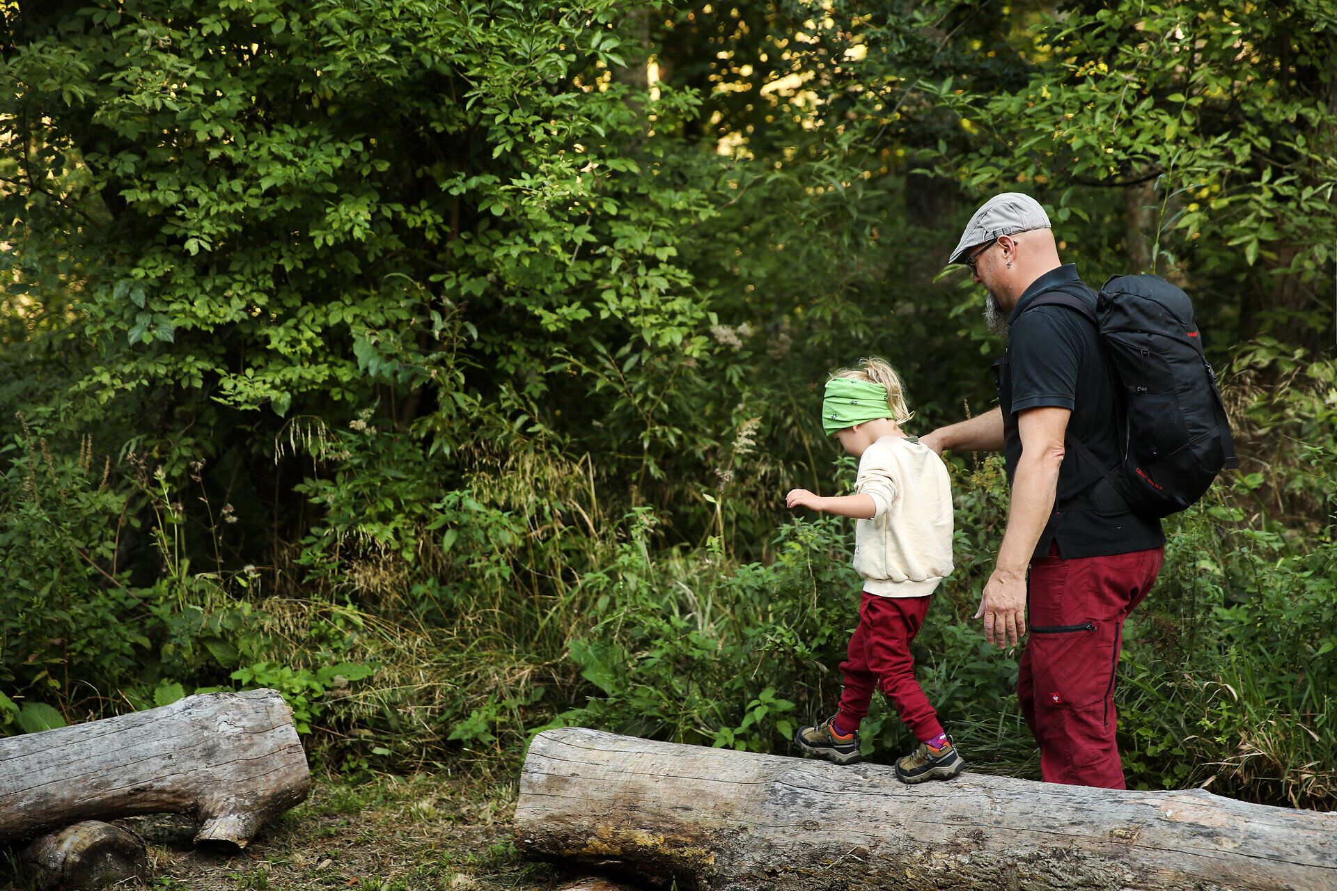 Ein Mann hält ein Kind an der Hand während dieses über einen Baumstamm balanciert. 
